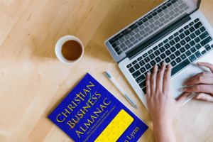 Hands typing on a laptop with a cup of coffee and a copy of Christian Business Almanac